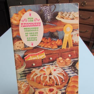 May include: A vintage cookbook titled The Fleischmann Treasury of Yeast Baking, featuring a colorful cover illustration of various baked goods, including a glazed ring cake, rolls, and pastries, with a yellow award ribbon and blue checkered tablecloth.