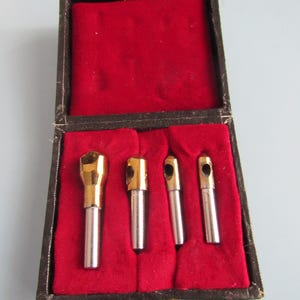 May include: A vintage set of four metal tools with gold-colored tops and silver shafts, displayed in a dark brown box lined with red velvet. The box is open, revealing the tools, and has a gold-colored clasp.