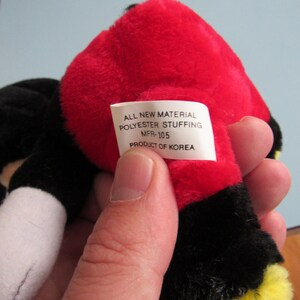 May include: A plush toy with a red head, black ears, and a black body. The toy has a white tag with the text "ALL NEW MATERIAL POLYESTER STUFFING MFR-105 PRODUCT OF KOREA".