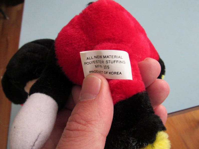 May include: A plush toy with a red head, black ears, and a black body. The toy has a white tag with the text "ALL NEW MATERIAL POLYESTER STUFFING MFR-105 PRODUCT OF KOREA".