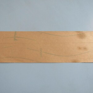 Op de afbeelding: Een rechthoekig stuk bruin papier met vage groene lijnen en tekst. Het papier heeft kreukels en vlekken, wat duidt op ouderdom of gebruik. De achtergrond is effen lichtblauw.