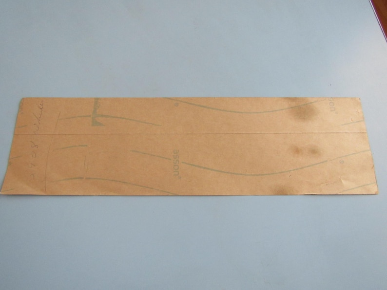 Op de afbeelding: Een rechthoekig stuk bruin papier met vage groene lijnen en tekst. Het papier heeft kreukels en vlekken, wat duidt op ouderdom of gebruik. De achtergrond is effen lichtblauw.