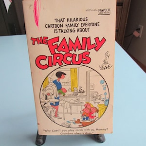 May include: A vintage paperback book titled "The Family Circus" by Bil Keane. The cover features a cartoon illustration of a family scene with text that reads "That Hilarious Cartoon Family Everyone Is Talking About". The book is propped up on a small black stand.