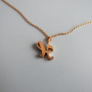 May include: A gold-colored necklace with a delicate chain. The pendant is a cursive letter "K". The necklace is displayed on a light gray background. The chain and pendant appear to be made of metal.
