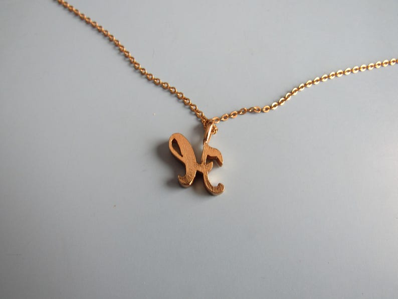 May include: A gold-colored necklace with a delicate chain. The pendant is a cursive letter "K". The necklace is displayed on a light gray background. The chain and pendant appear to be made of metal.