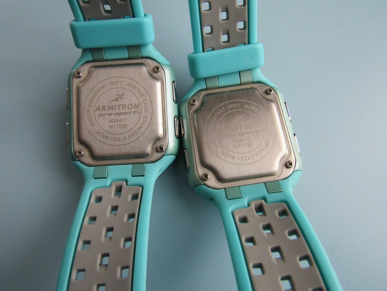 May include: Two Armitron Pro Sport digital watches with teal and gray straps. The stainless steel backs display the brand name, model number, and water resistance details. The watches have an acrylic case and are water resistant to 165 feet.