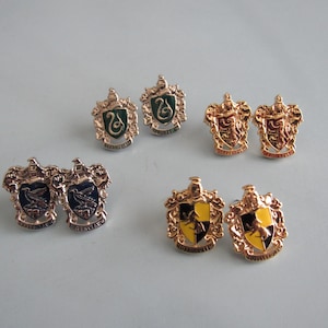 May include: Four pairs of gold-toned metal pins shaped like heraldic shields. Each pair features a different colored enamel center: green with a serpent, red with a lion, blue with an eagle, and yellow with a badger, all with ornate metal borders.