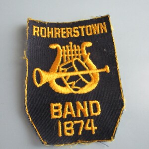 May include: A black embroidered patch with gold text and a musical instrument design. The text reads "ROHRERSTOWN BAND 1874". The patch has a unique shape and a gold border.
