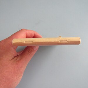 May include: A light brown, rectangular object, possibly a package or a wrapped item, held in a hand. The object has a seam or closure visible on its side. The background is a solid, light blue-gray color.
