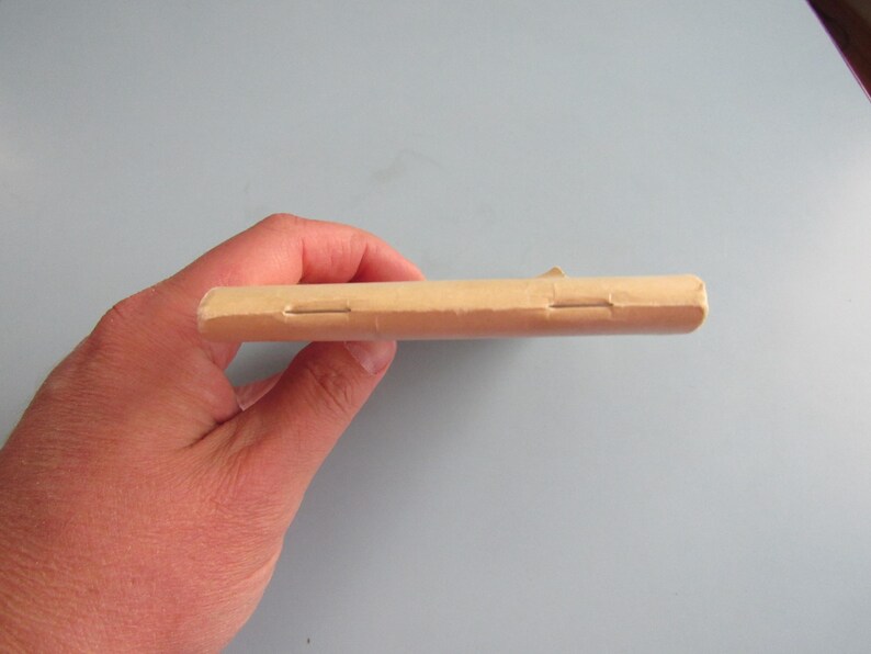 May include: A light brown, rectangular object, possibly a package or a wrapped item, held in a hand. The object has a seam or closure visible on its side. The background is a solid, light blue-gray color.