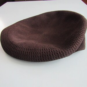 May include: A brown knitted cap with a rounded crown and a short, curved brim. The cap is made of a textured fabric, and the colour is a deep, earthy brown. The cap is on a white surface.