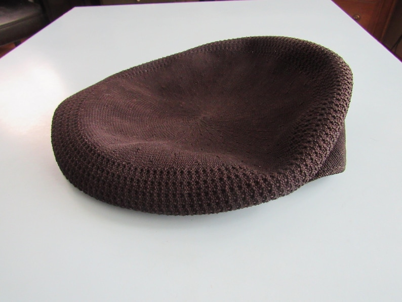 May include: A brown knitted cap with a rounded crown and a short, curved brim. The cap is made of a textured fabric, and the colour is a deep, earthy brown. The cap is on a white surface.