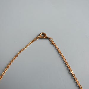 May include: A gold-colored necklace chain with a lobster clasp. The chain is made of small, interconnected links. The clasp is attached to a small ring. The background is a solid, light gray color.