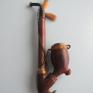 May include: A vintage tobacco pipe with a dark brown bowl and stem, accented with brass fittings and a decorative tassel. The pipe has a curved design and a long, slender stem. The overall length is approximately 12 inches.