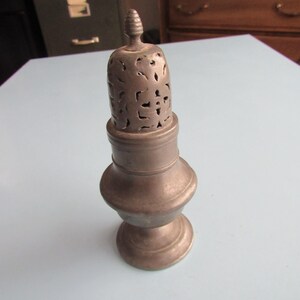 May include: A metallic sugar shaker with a dull gray finish, featuring a bulbous body, a flared base, and a removable domed lid with intricate cut-out patterns and a small, pointed finial at the very top of the shaker's lid structure.