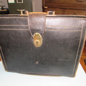 May include: A rectangular black leather briefcase featuring a textured surface, visible stitching along the edges, and a central vertical strap secured by a gold-toned metal latch. The top includes a sturdy handle with metal hardware attachments.