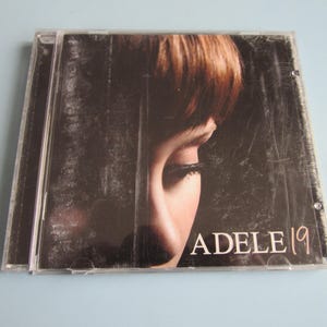 May include: A clear plastic CD jewel case featuring the album cover for Adele 19. The cover art displays a close-up of a person's face in profile, partially obscured by dark, textured vertical lines. The text ADELE 19 is printed in white.