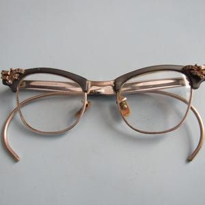 May include: Vintage eyeglasses with a black upper frame and gold-colored lower frame and arms. The upper frame has decorative embellishments on the corners. The lenses are clear. The left arm is detached. The glasses are set against a light blue background.