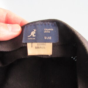 May include: A close-up of a black Kangol hat. The interior label shows the Kangol logo, the word "BLUE", and the size "SMALL". The label also includes the text "FOUNDED 38.83".