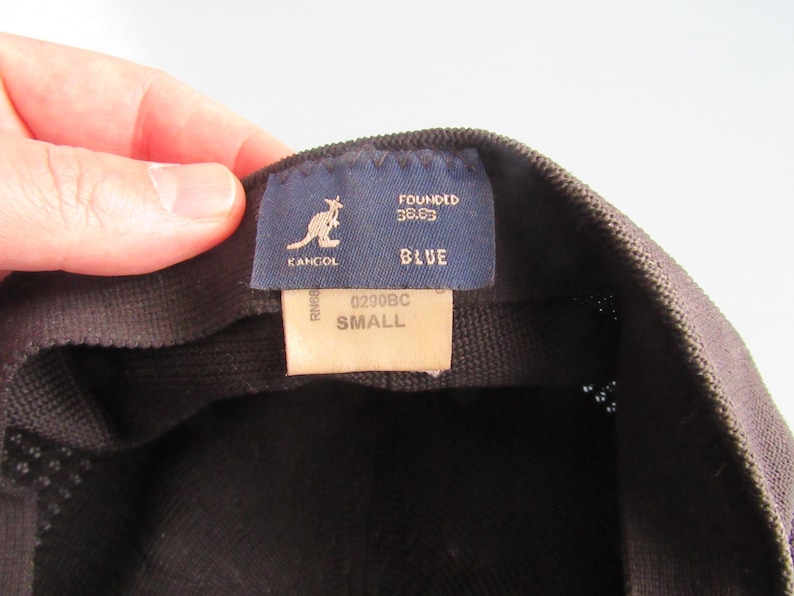 May include: A close-up of a black Kangol hat. The interior label shows the Kangol logo, the word "BLUE", and the size "SMALL". The label also includes the text "FOUNDED 38.83".