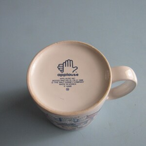 May include: A white ceramic mug viewed from the bottom, featuring a blue logo of a hand and the text Applause Inc. Woodland Hills, CA 1988 The Walt Disney Company Made in Korea #14666. The mug has a curved handle extending from the right side.