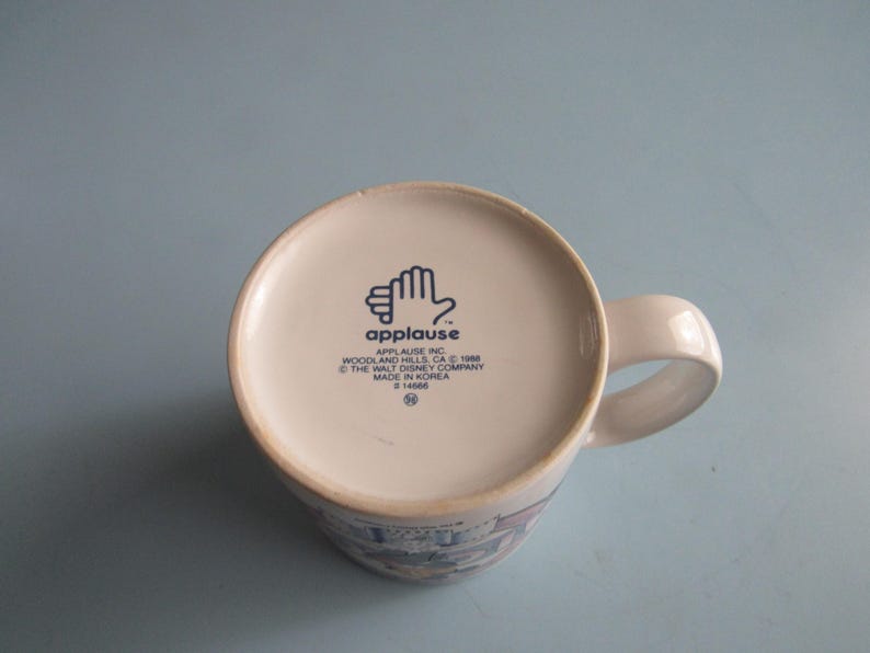 May include: A white ceramic mug viewed from the bottom, featuring a blue logo of a hand and the text Applause Inc. Woodland Hills, CA 1988 The Walt Disney Company Made in Korea #14666. The mug has a curved handle extending from the right side.