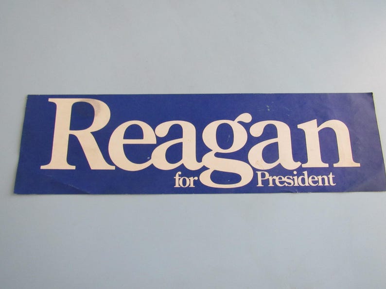 Op de afbeelding: Een blauwe rechthoekige sticker met de witte tekst "Reagan for President". Het woord "Reagan" staat in een groot, vetgedrukt lettertype en "for President" staat eronder in een kleiner lettertype.