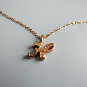 May include: A gold-colored necklace with a cursive letter "H" pendant. The pendant is embellished with small, clear crystals. The chain is a delicate link style, and the necklace is displayed against a light background.
