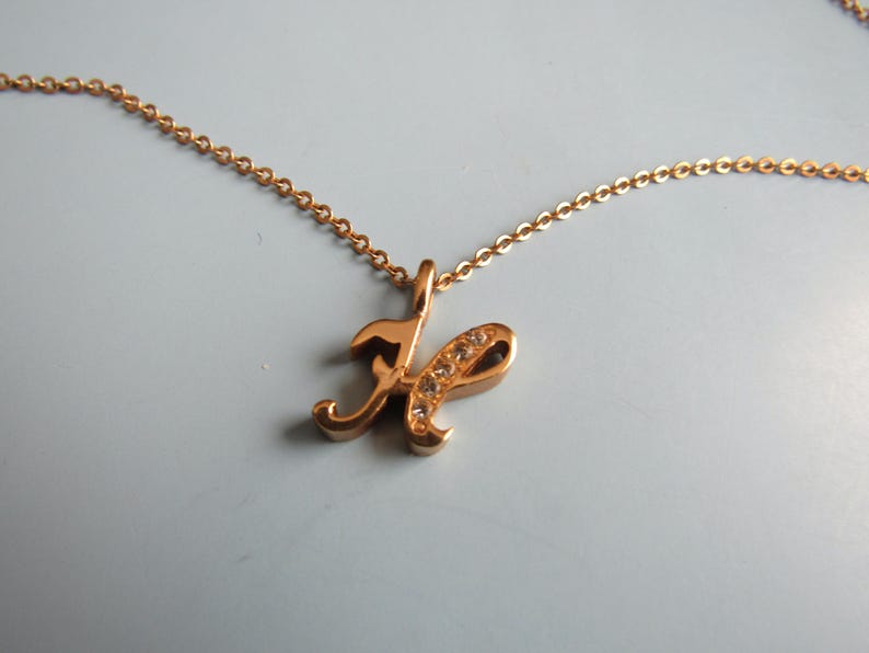 May include: A gold-colored necklace with a cursive letter "H" pendant. The pendant is embellished with small, clear crystals. The chain is a delicate link style, and the necklace is displayed against a light background.