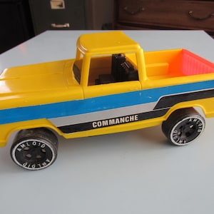 May include: A vintage yellow toy truck with black wheels and the word "COMANCHE" printed on the side. The truck has blue and silver stripes and a red bed. A classic toy vehicle.