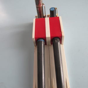 May include: A vintage pen holder with a red and white rectangular top section and two black cylindrical compartments. Two pens are visible, one silver and one black with the number "2/3" printed on it. The holder is made of plastic and has a retro design.