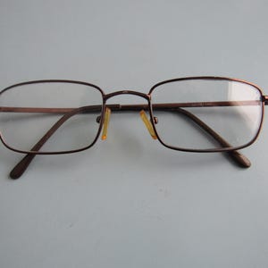 May include: Brown rectangular eyeglasses with clear lenses. The glasses have a thin, metal frame and small, yellow nose pads. The temples are a darker brown. The glasses are designed for vision correction.