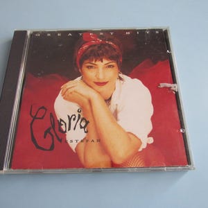 May include: A CD case with a red and white background. The cover features a woman with red hair wearing a white shirt and a red headband. The text "Gloria Estefan Greatest Hits" is printed on the cover.