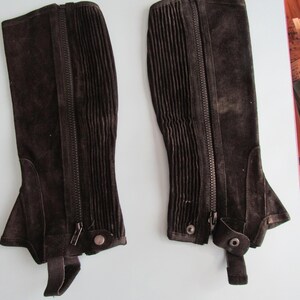 May include: A pair of black suede half chaps featuring a vertical zipper closure, a ribbed panel along the side, and a bottom strap with a snap button fastener. These equestrian leg guards are designed to cover the lower leg from ankle to knee.