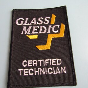 May include: A rectangular, brown fabric patch with the words "GLASS MEDIC" in white and "CERTIFIED TECHNICIAN" in white. The patch has a yellow cross design and a black border.