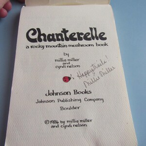 May include: Open book titled "Chanterelle, a rocky mountain mushroom book" by Millie Miller and Cyndi Nelson. The cover is off-white with black text and a red ladybug graphic. Handwritten note says "Happy trails! Millie Miller". Published by Johnson Books in 1986.