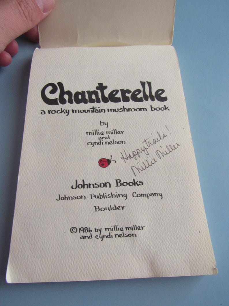 May include: Open book titled "Chanterelle, a rocky mountain mushroom book" by Millie Miller and Cyndi Nelson. The cover is off-white with black text and a red ladybug graphic. Handwritten note says "Happy trails! Millie Miller". Published by Johnson Books in 1986.