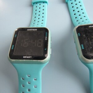May include: Two Armitron digital watches with turquoise bands. The watch faces are black with white text displaying the time 15:48. The watch bands have a series of rectangular perforations. The watch cases are square with rounded corners.
