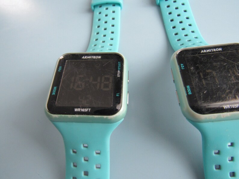 May include: Two Armitron digital watches with turquoise bands. The watch faces are black with white text displaying the time 15:48. The watch bands have a series of rectangular perforations. The watch cases are square with rounded corners.