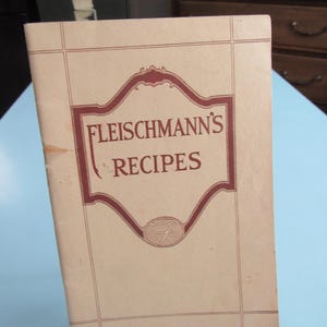 May include: A vintage softcover recipe booklet titled Fleischmann's Recipes. The cover is light beige with a dark red decorative border and text. A stylized letter F is enclosed in an oval at the bottom center. The booklet has a vertical orientation.