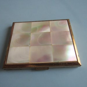 May include: A square compact mirror featuring a grid of nine iridescent mother-of-pearl tiles set within a metallic gold-toned frame. The surface displays a soft, pearlescent sheen with subtle pink and green hues across the tiles.