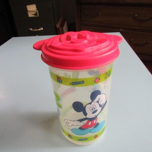 May include: A clear plastic tumbler featuring a graphic of Mickey Mouse and a bright pink lid molded with the shape of Mickey Mouse ears. The cup has a green band around the top and bottom with small illustrations of apples and juice cartons.