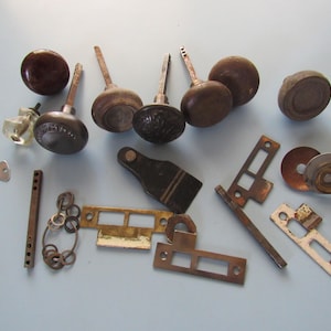 May include: An assortment of vintage hardware components, including several brown wooden door knobs, metal latches, a key, and a small glass bottle. The hardware pieces are arranged on a light blue surface.