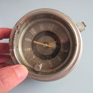 May include: Vintage round clock with a silver-colored bezel and a dark gray face. The clock features gold-colored hands and numerals. The clock is held by a hand. The clock is an antique.