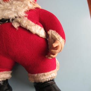 May include: A vintage Santa Claus doll, featuring a red felt suit trimmed with white faux fur. The doll has a white beard, a rosy face, and black boots. The doll's arms are bent, and the hands are visible.