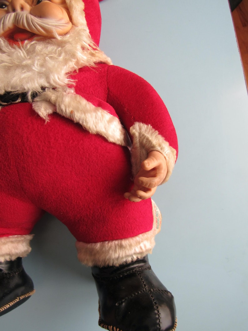 May include: A vintage Santa Claus doll, featuring a red felt suit trimmed with white faux fur. The doll has a white beard, a rosy face, and black boots. The doll's arms are bent, and the hands are visible.