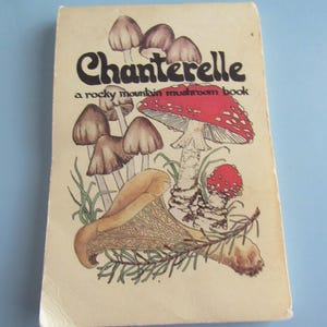 May include: A vintage book titled "Chanterelle" with a mushroom illustration. The cover features a red and white spotted mushroom, brown mushrooms, and the text "a rocky mountain mushroom book". The book has a cream-colored cover.