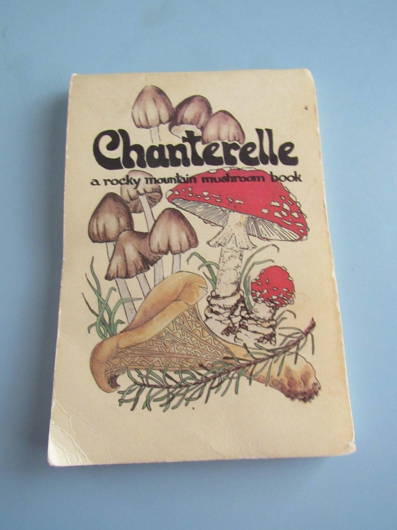 May include: A vintage book titled "Chanterelle" with a mushroom illustration. The cover features a red and white spotted mushroom, brown mushrooms, and the text "a rocky mountain mushroom book". The book has a cream-colored cover.