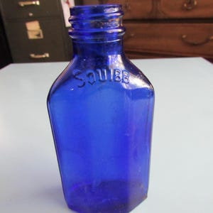 May include: A cobalt blue glass bottle with a rounded top and a squared bottom. The bottle has the word "SQUIBB" embossed on the front.