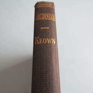 May include: A brown hardcover book spine with a textured fabric finish. The title "MECHANISM" is printed in gold lettering near the top, followed by a horizontal gold line and the author name "KEOWN" in gold lettering centered below.